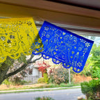 Three colorful papel picado banners in yellow, blue, and pink hanging across a window, with a suburban street and trees visible outside.