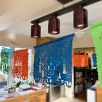 Colorful Mexican papel picado banner decorations hanging from a ceiling track light in a home interior setting.