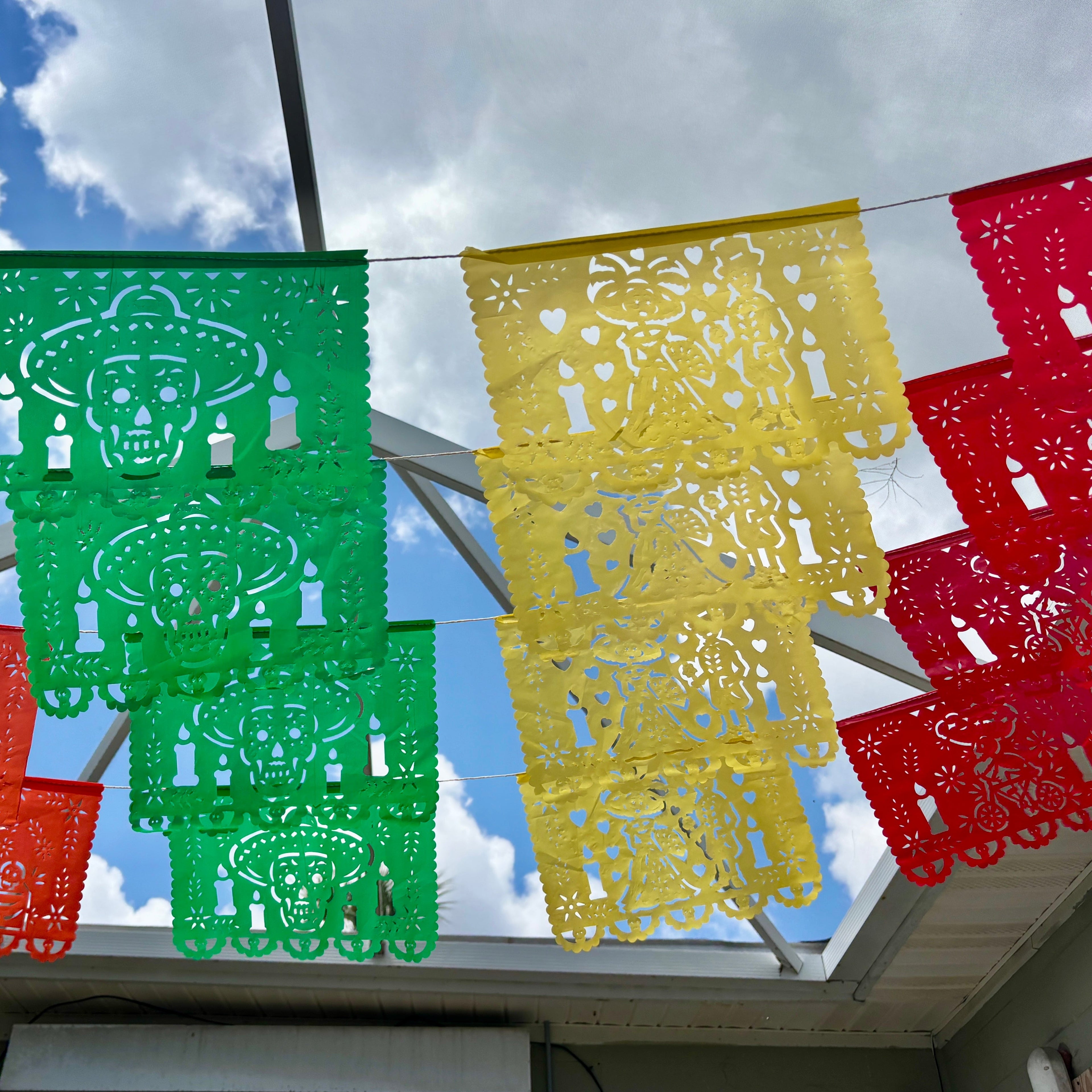 Colorful Mexican papel picado banners hanging in rows under a cloudy blue sky, with green, yellow, red, orange, and blue panels strung across a patio structure.