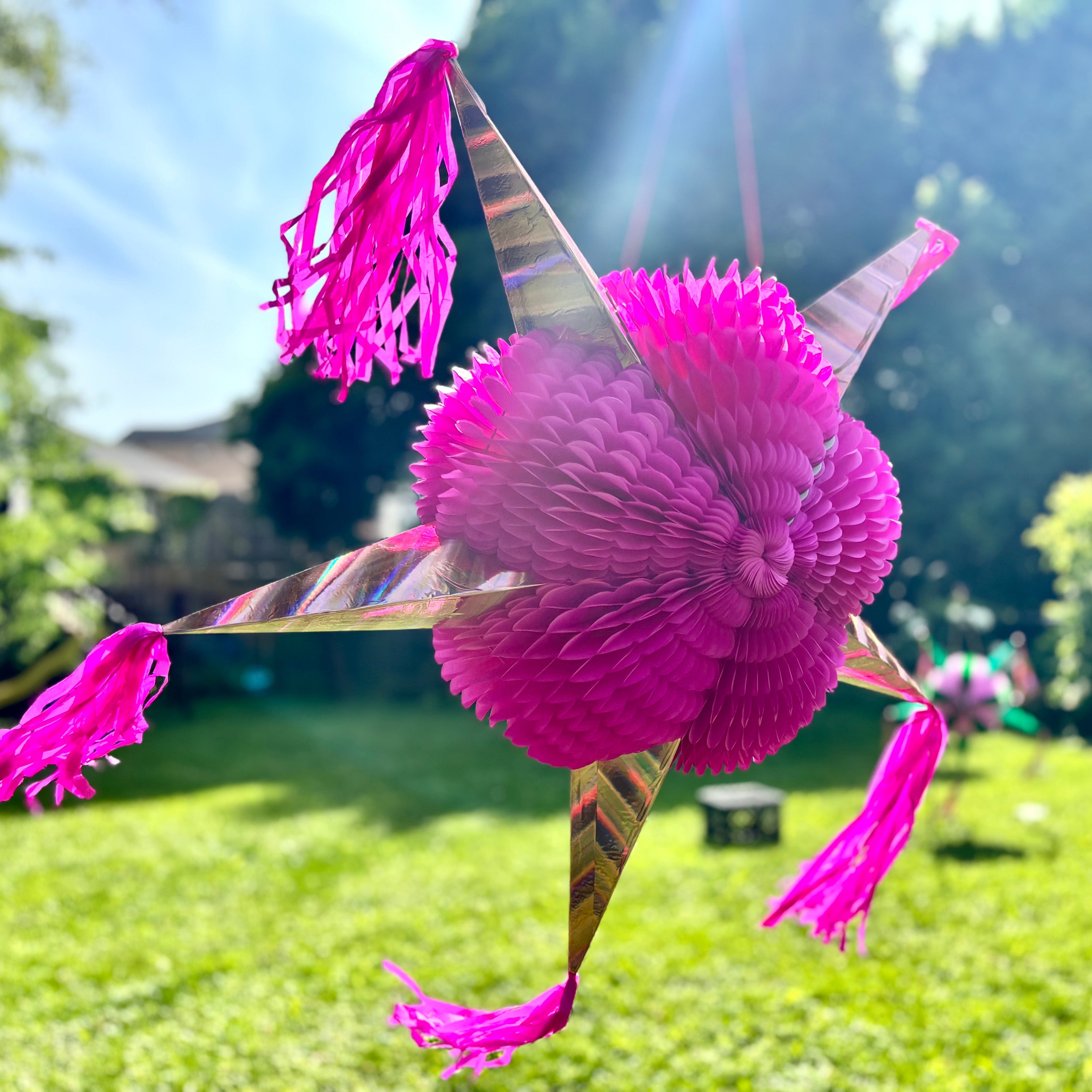 A bright pink star-shaped paper piñata hanging outdoors in a sunny backyard with green grass and trees in the background.