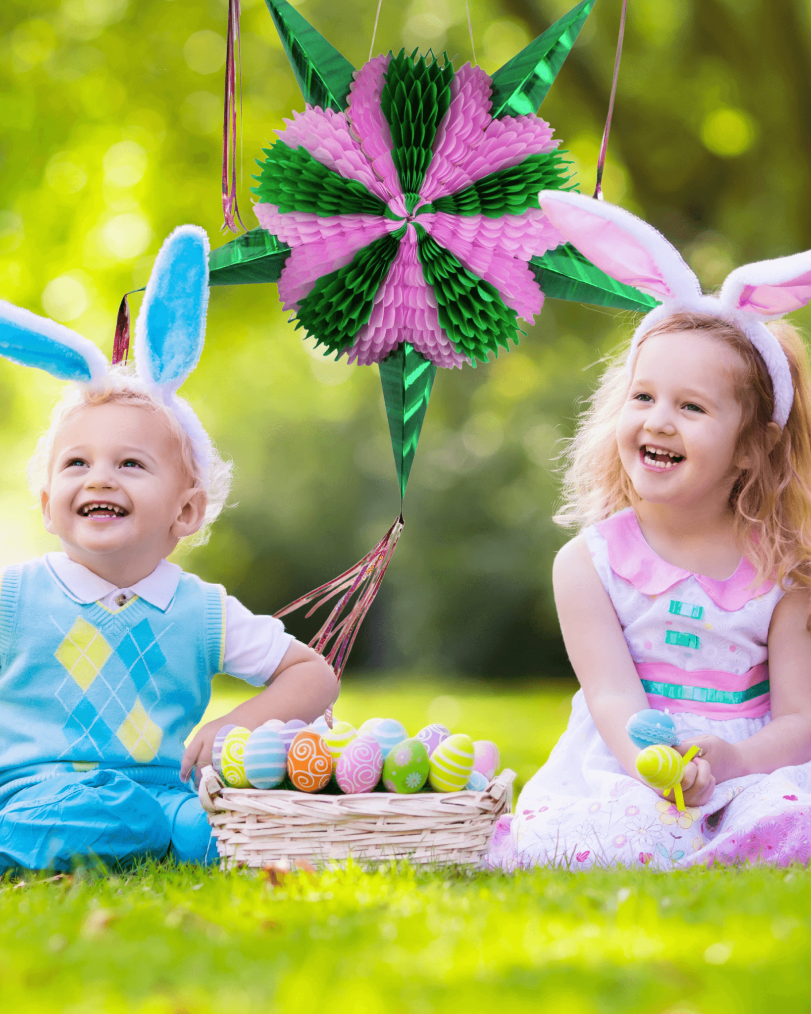 Two children wearing bunny ears sitting on grass with Easter eggs and a decorative pinata.